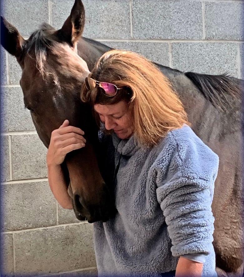 A woman in a blue sweater affectionately resting her head against a brown horse in a stable setting.