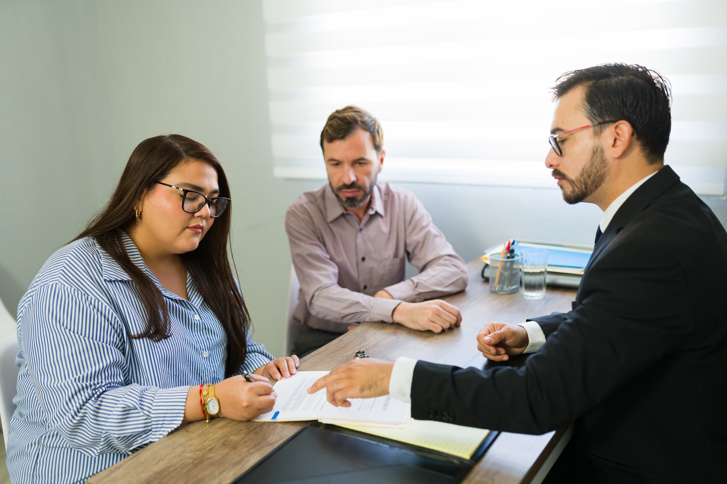 Woman signing legal agreement in a lawyer's office, husband sitting next to her during a conciliation meeting or separation process