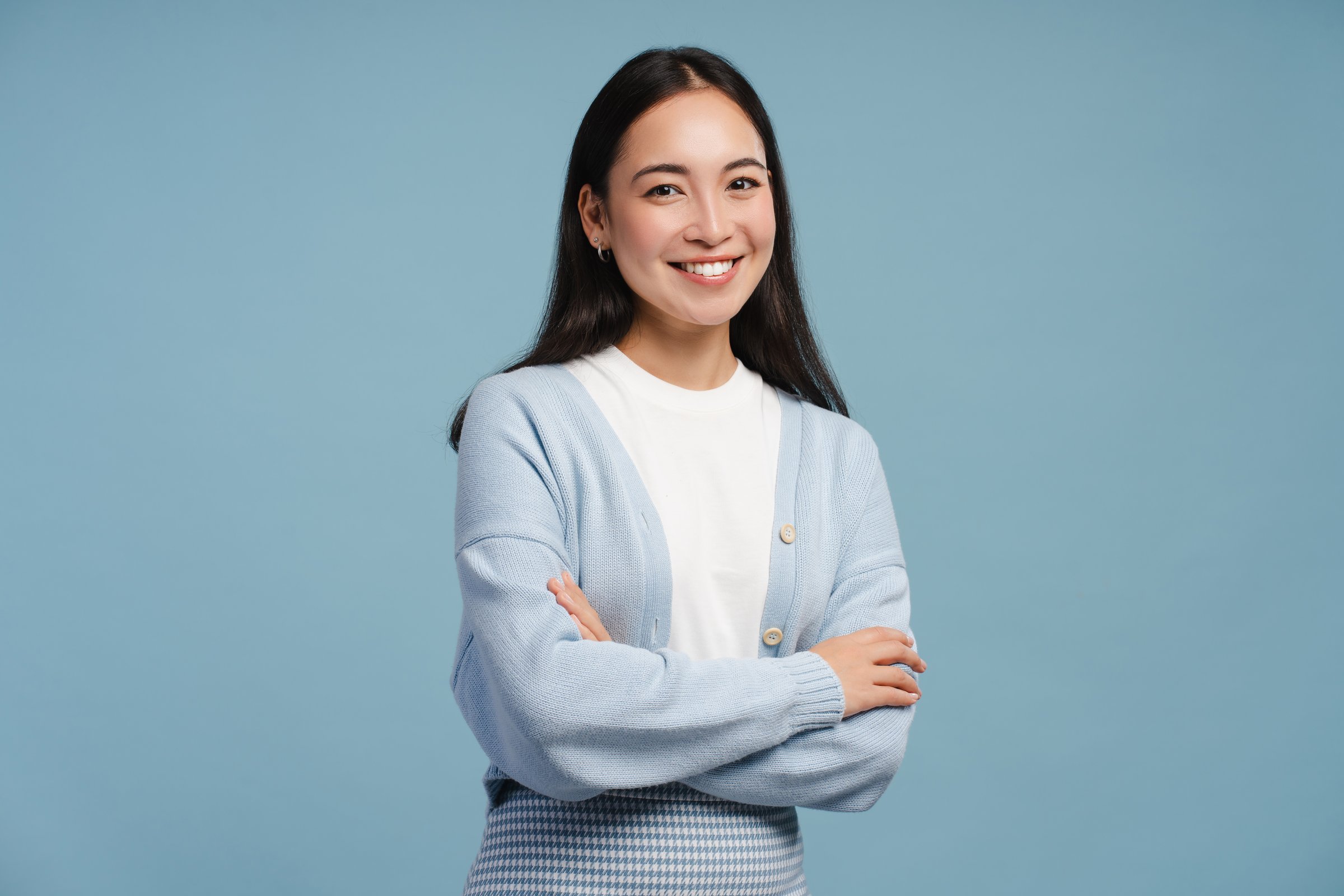 Smiling, young Asian woman with crossed arms, looking at camera, standing isolated on blue background, posing in studio. Advertisement concept