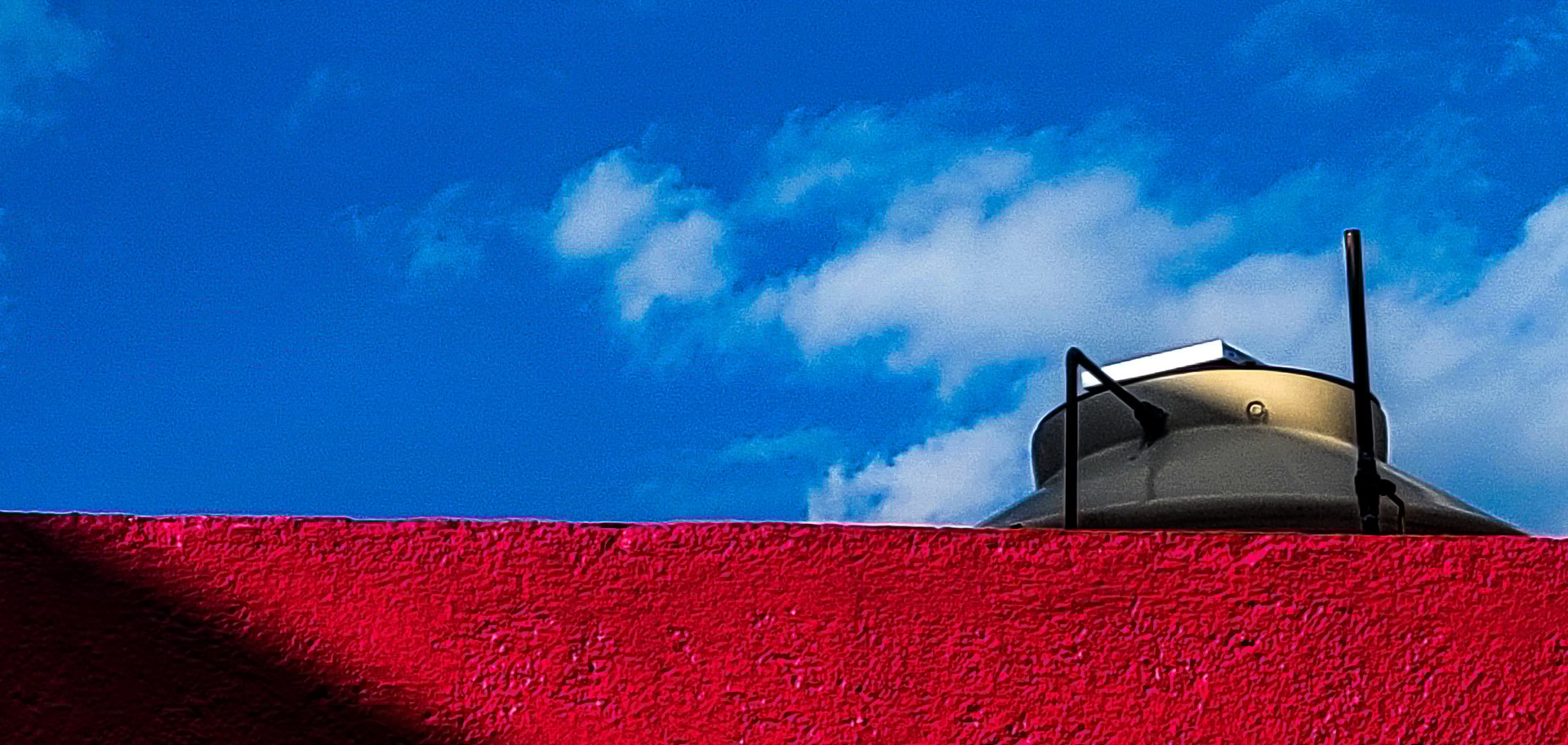 Red wall and rooftop water tank under a bright blue sky with scattered clouds.