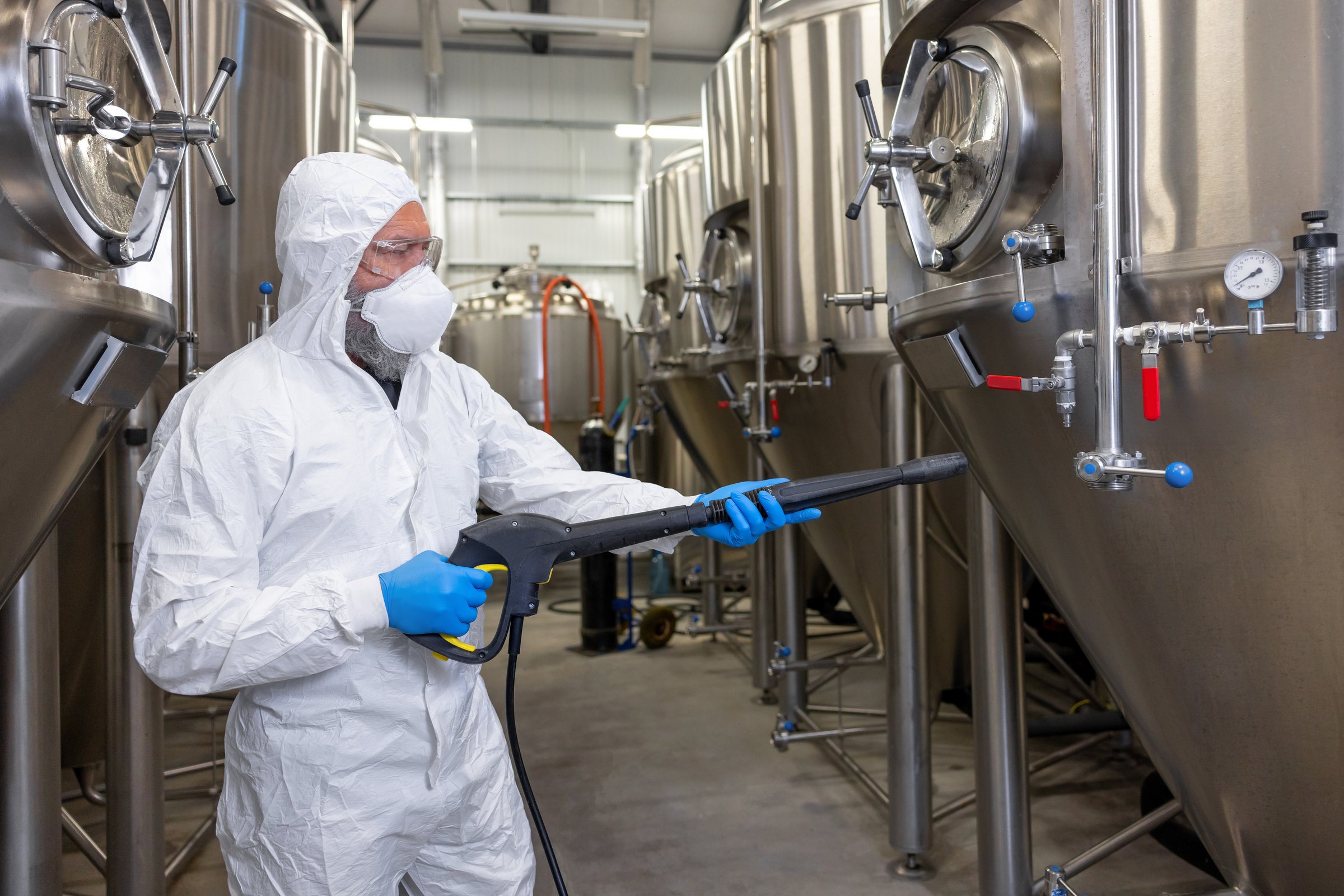 Focused factory worker in a protective mask and nitrile gloves aiming the pressure washer at the stainless steel fermentation tank