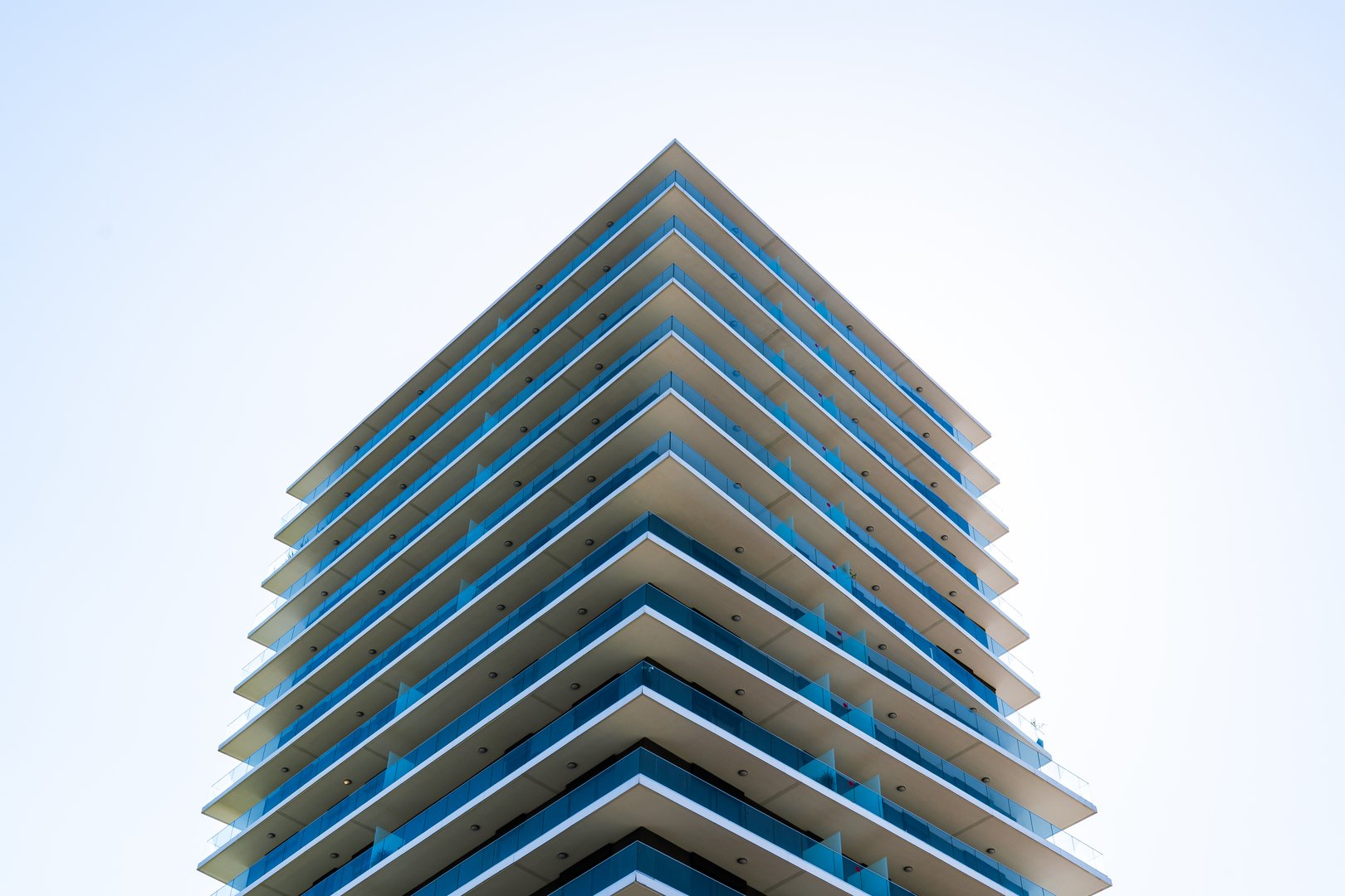 This image shows a contemporary residential building featuring a sleek façade with alternating balconies and glass railings. The sharp geometry and minimal design emphasize both functionality and modern aesthetics. Set against a bright sky, the building conveys elegance, comfort, and urban living.