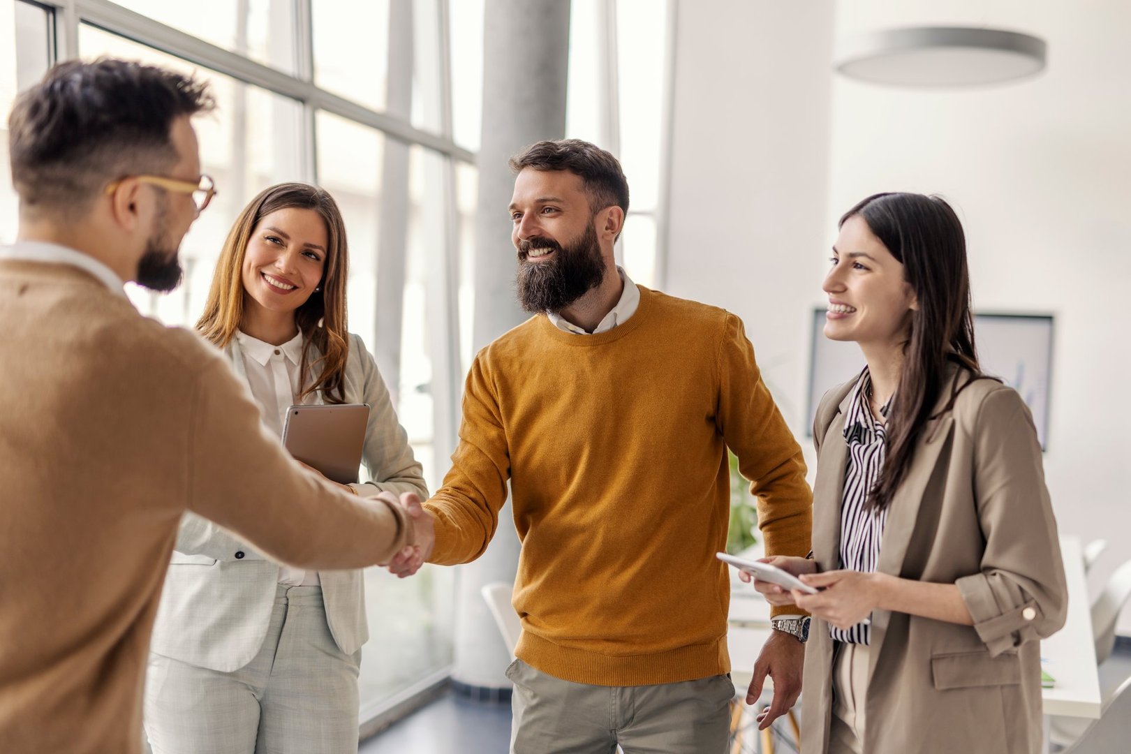A diverse group of business professionals, including men and women, collaborates in a modern, well-lit office. One man in a mustard sweater shakes hands with a colleague.