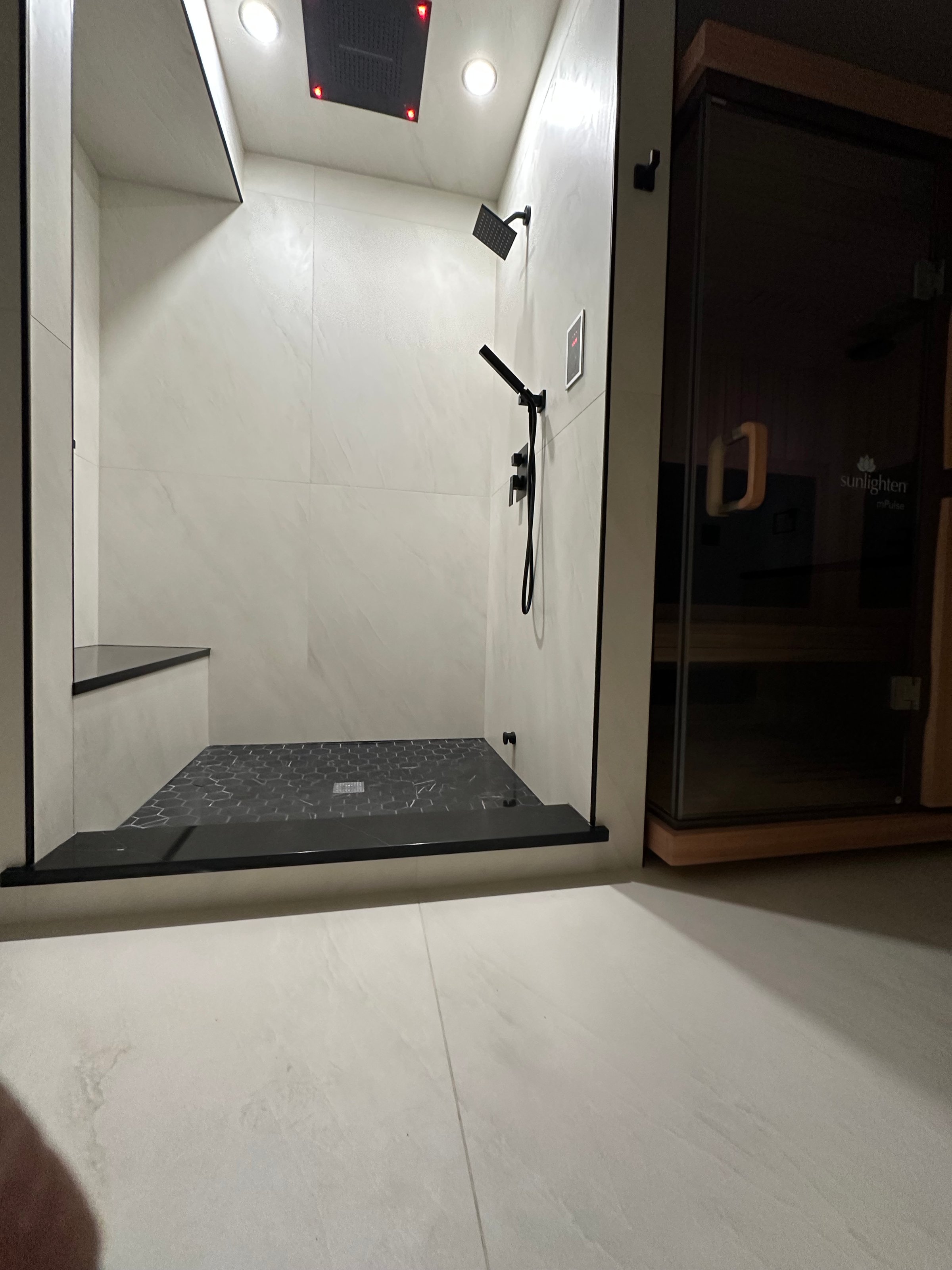 Modern bathroom with a large shower, black showerhead, and a wooden sauna room visible to the right.