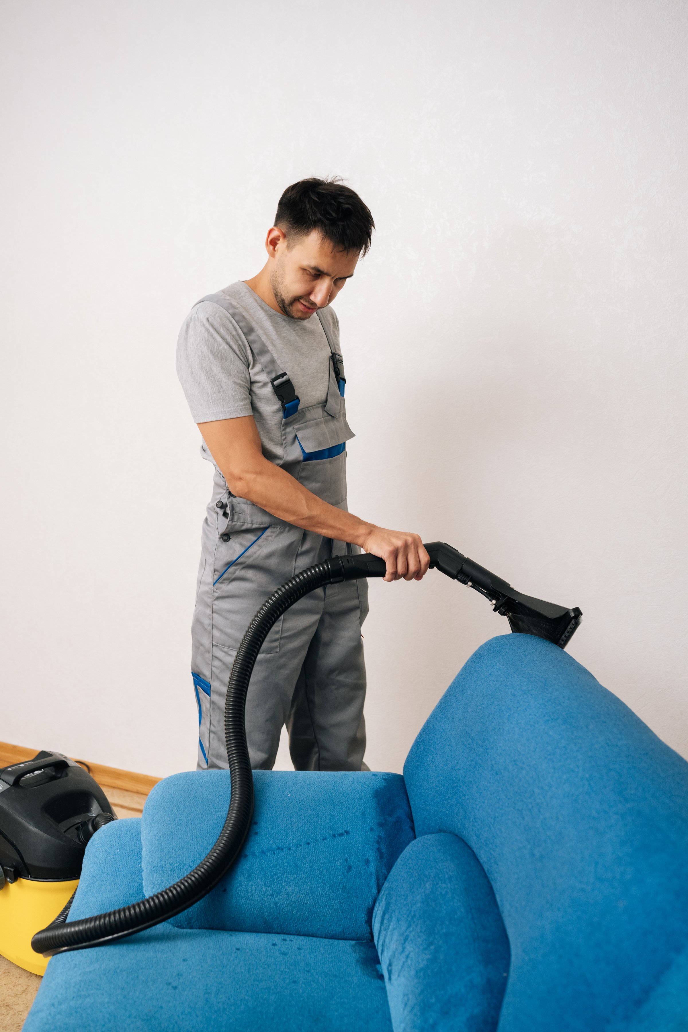 Vertical portrait of professional cleaning service worker removing dirt and stains from blue upholstered sofa using extraction washing vacuum cleaner in living room. Concept of household cleanliness.