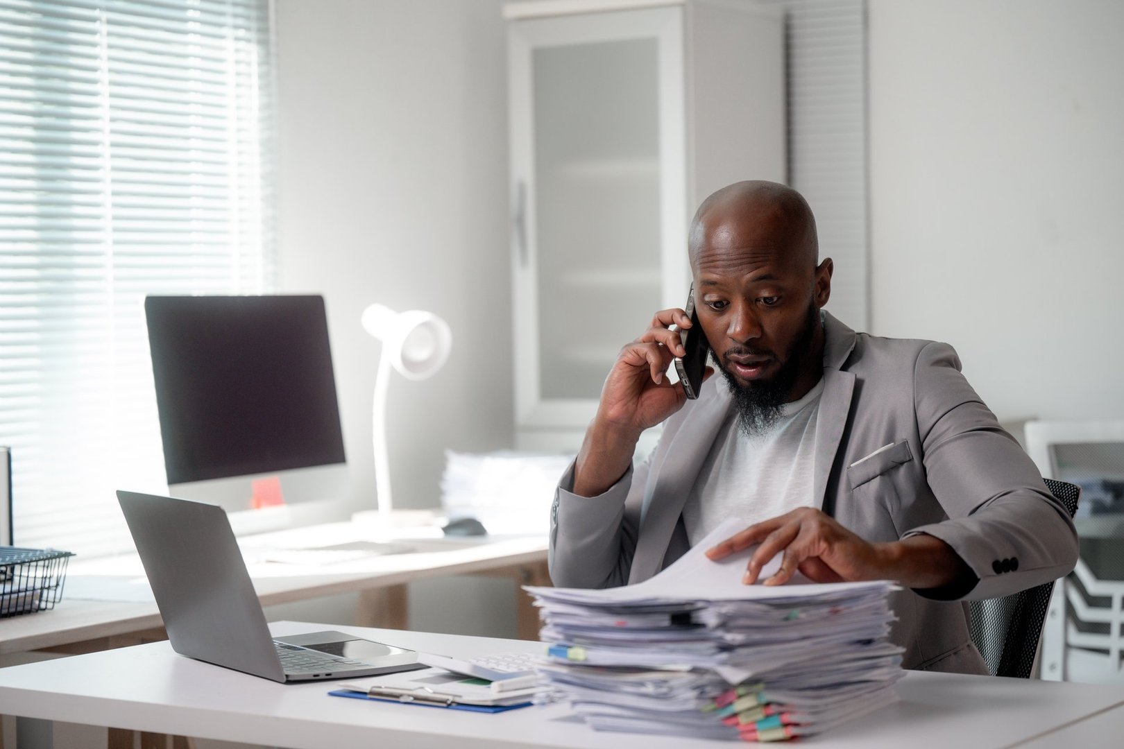 Stressed businessman having a phone call while searching through a pile of paperwork at his office desk