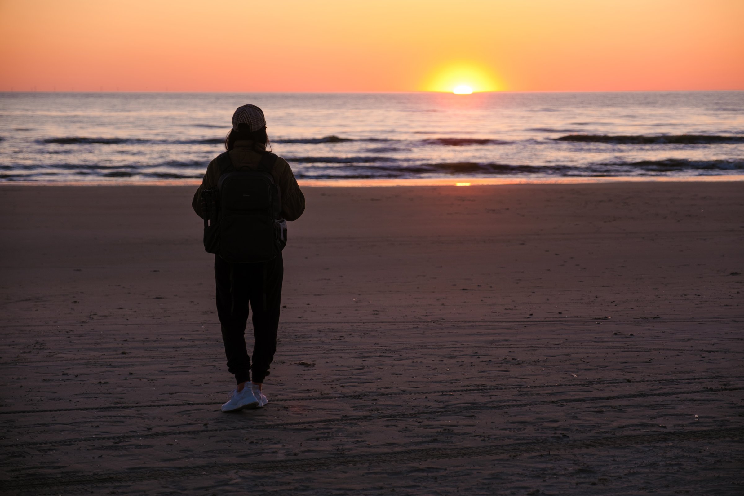 A serene evening unfolds as a person walks along a beautiful Danish beach during sunset. The vibrant orange sun dips below the horizon, reflecting on calm waters, Vejers Strand beach Denmark