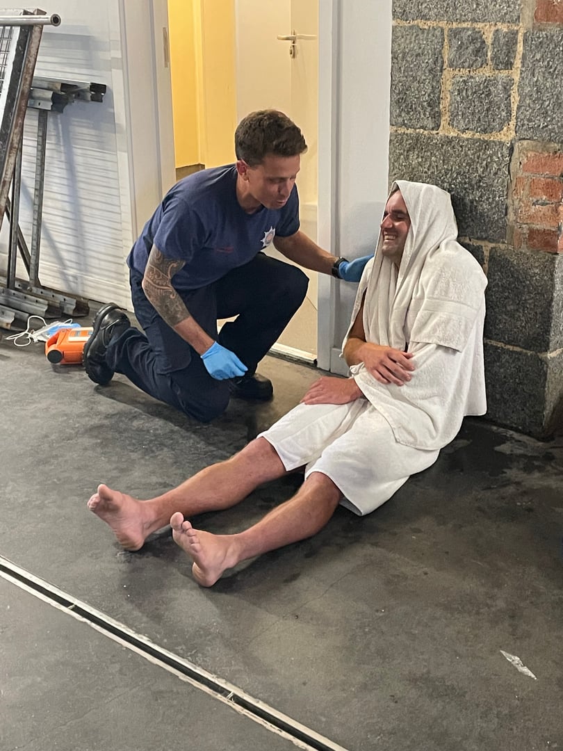 Paramedic assisting a seated, smiling man covered in a towel against a wall in a building hallway.