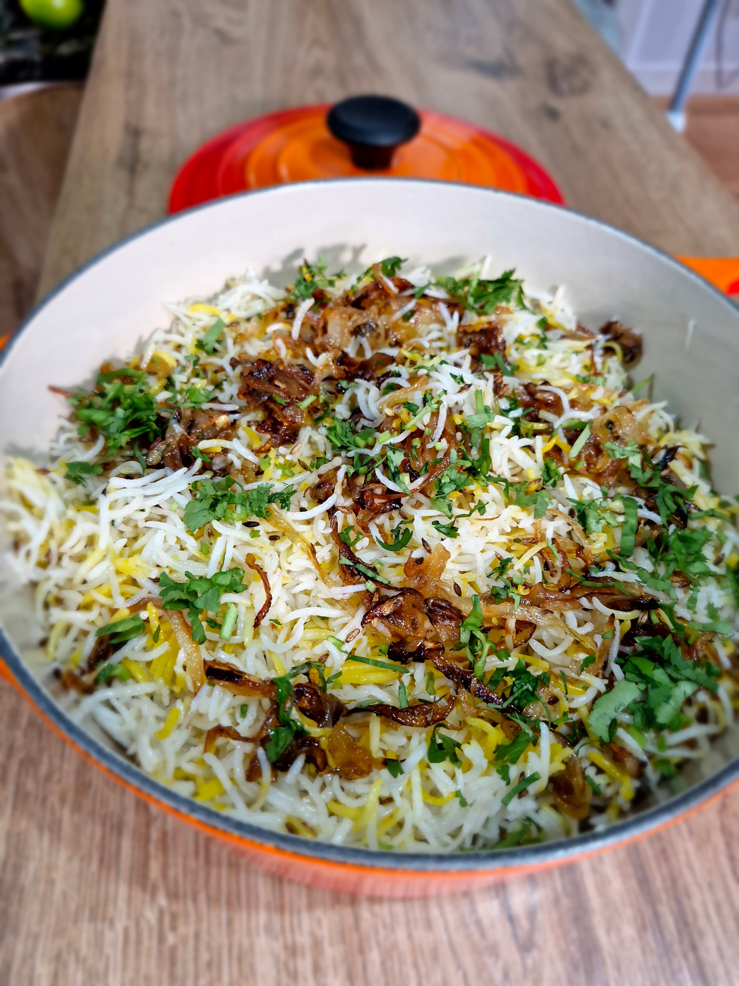 A pot of biryani with saffron rice, caramelized onions, and fresh herbs on a wooden table, next to an orange lid.