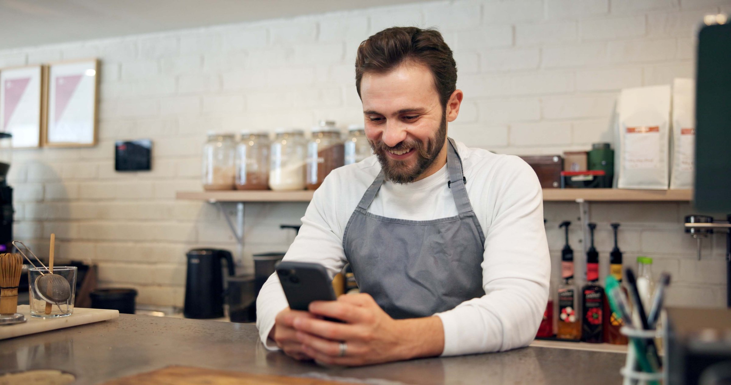 Happy man, barista and chatting with phone at cafe counter for small business, social media or review. Male person, bistro or employee with smile on mobile smartphone for online app at coffee shop