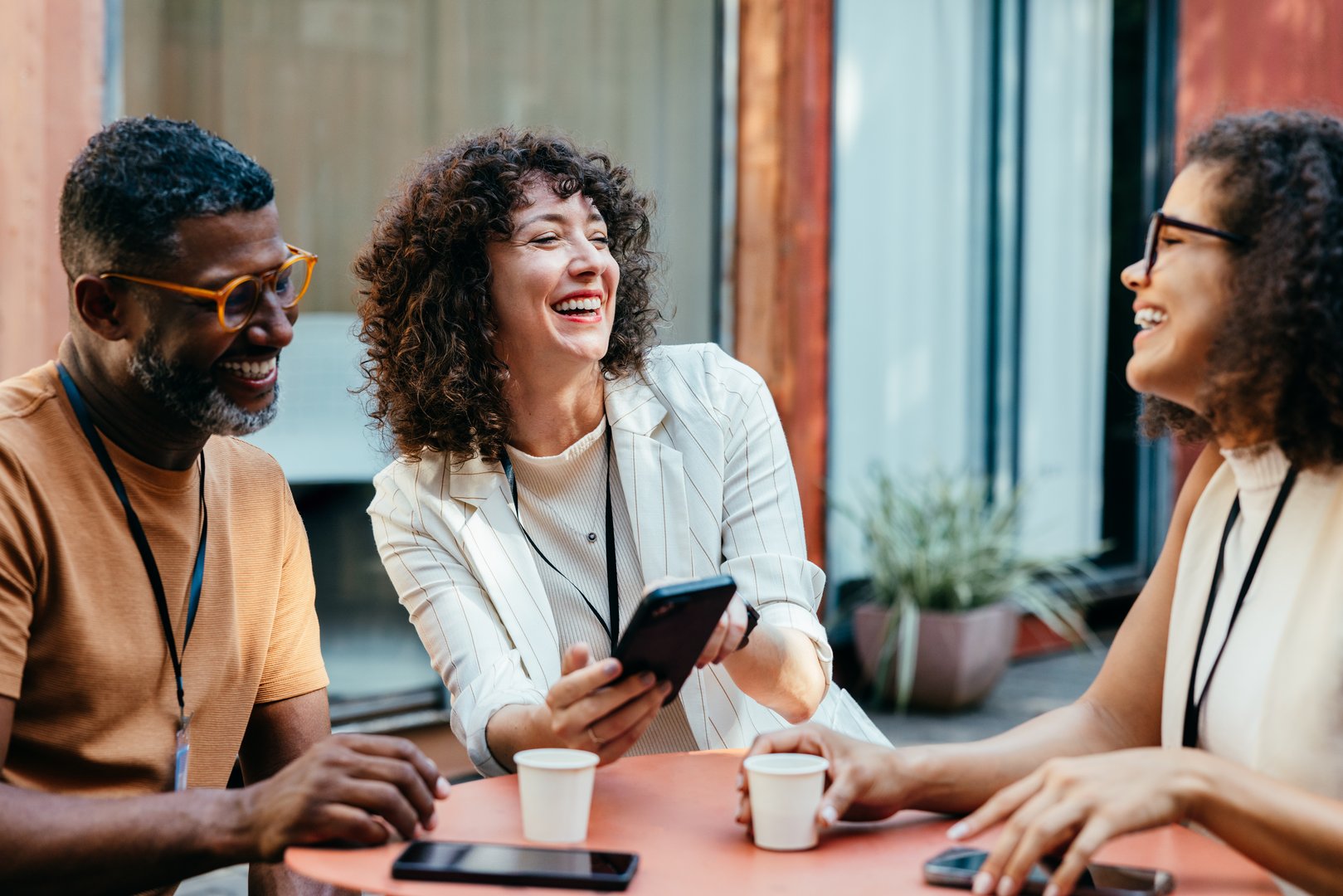 A group of coworkers from different backgrounds enjoying a cheerful coffee break together, gathered around an outdoor table, laughing over something on a phone.