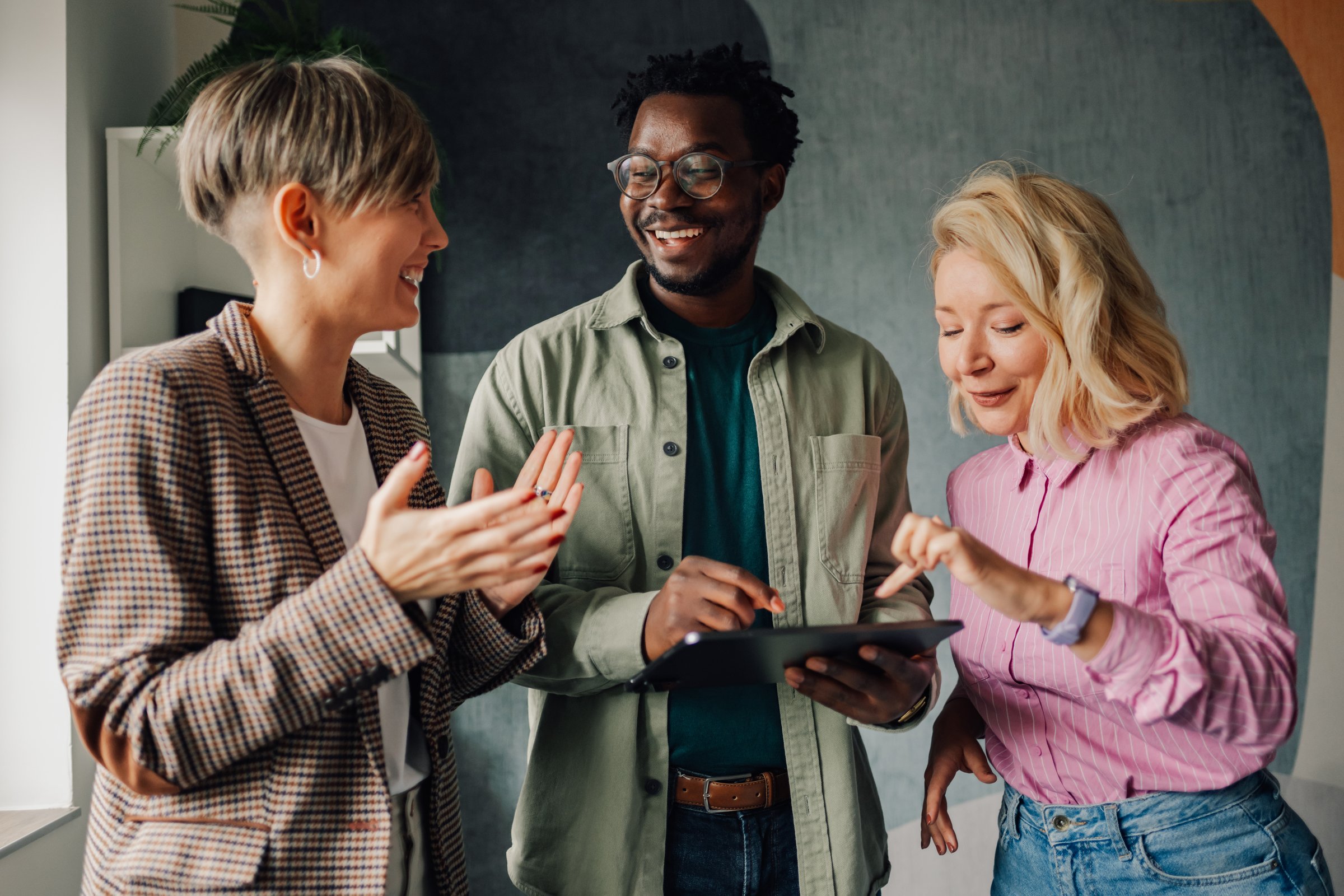 Three smiling colleagues are discussing a project, using a digital tablet and exchanging ideas in a modern office environment, showcasing teamwork and collaboration