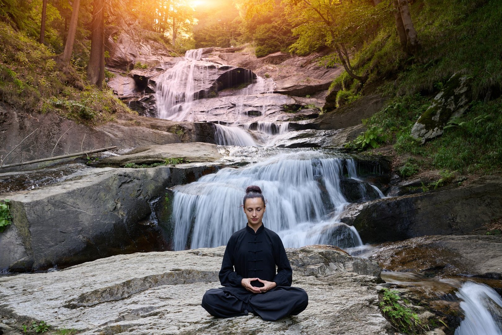 A woman sits in lotus pose practicing yoga under a tropical waterfall