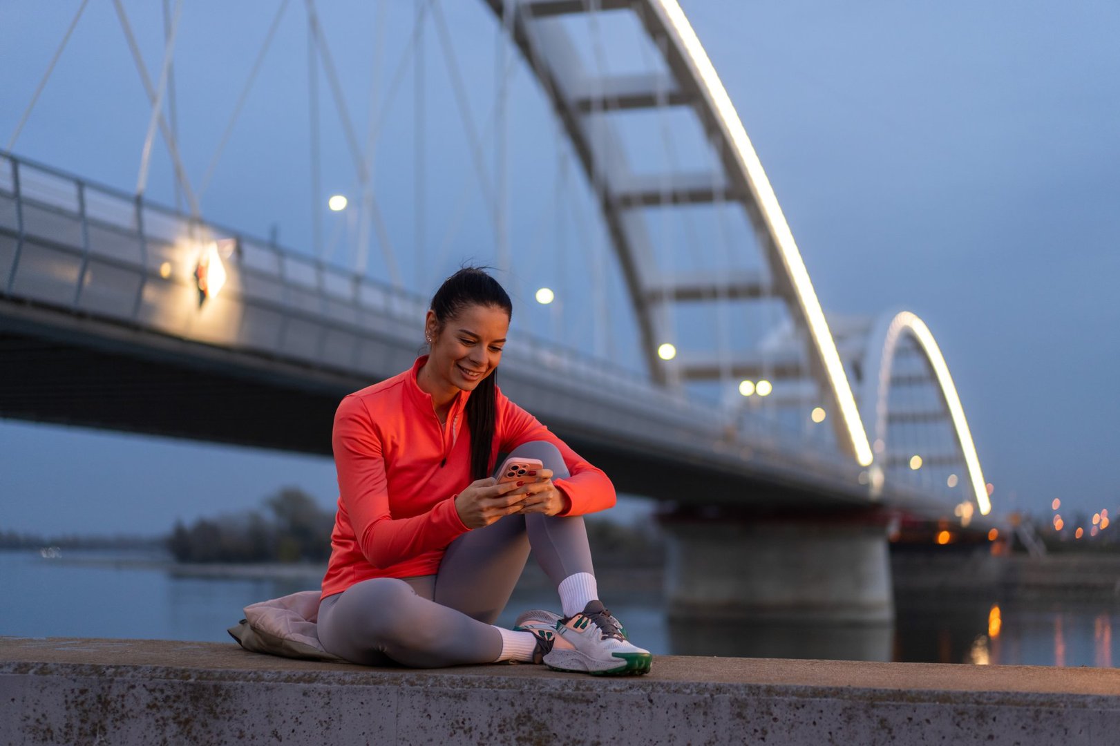 Young adult woman smiling using cell phone after a workout outdoors at sunset
