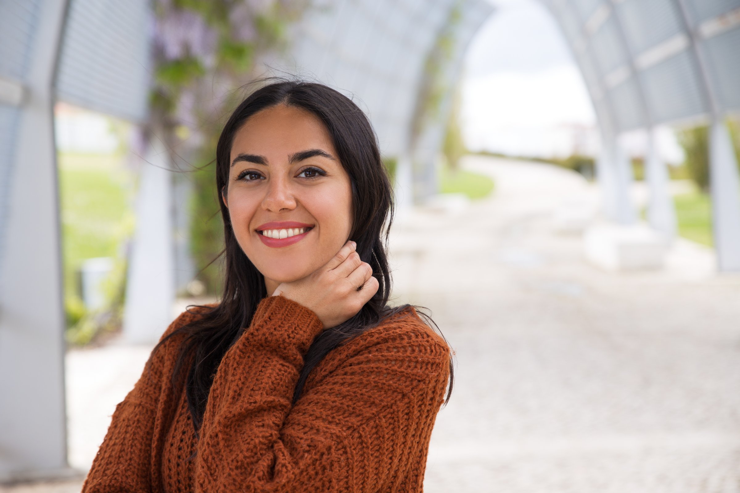 Woman in a brown sweater smiling outdoors in a covered walkway with a blurred background.