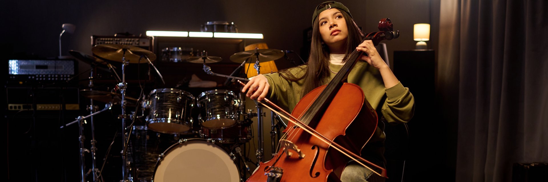 Latin girl playing cello in music studio, sitting in front of drum set and amplifiers, wearing casual clothing, focused expression, long dark hair visible