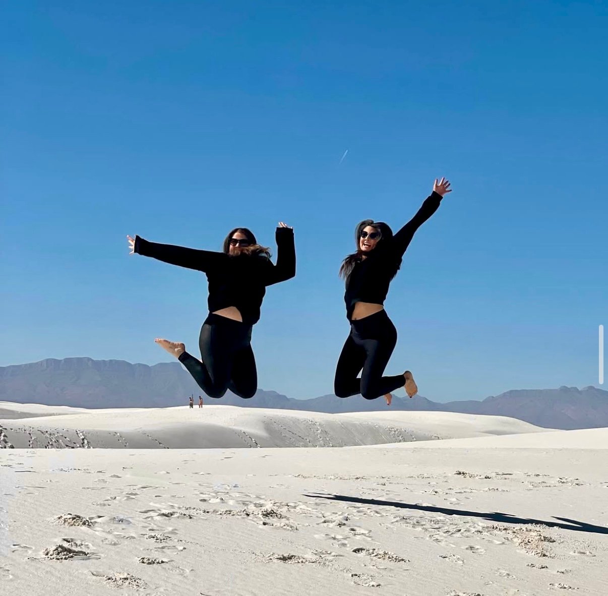 Two people in black outfits jumping on a sandy desert landscape with mountains in the background and a clear blue sky.