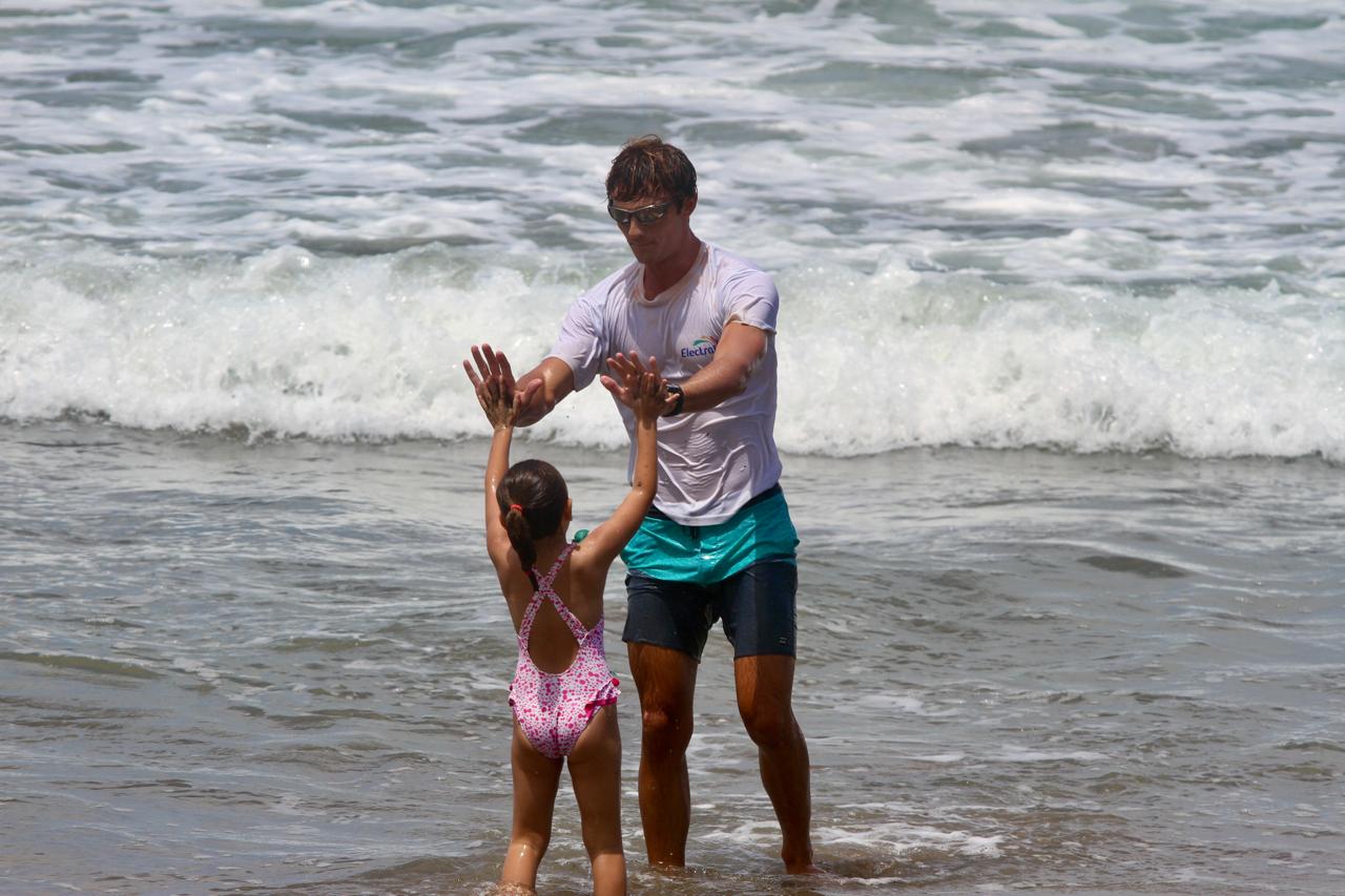 Man and young girl playing in ocean waves, holding hands. Man wears sunglasses, girl in pink swimsuit. Surf in background.