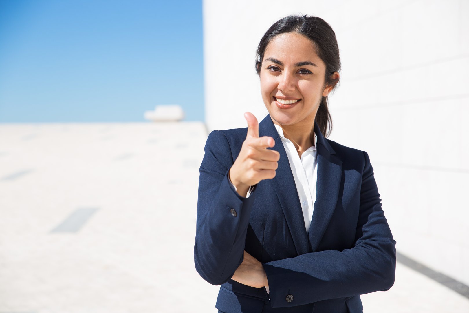 Smiling woman in a business suit pointing towards the camera, standing outside against a clear sky and modern architecture.