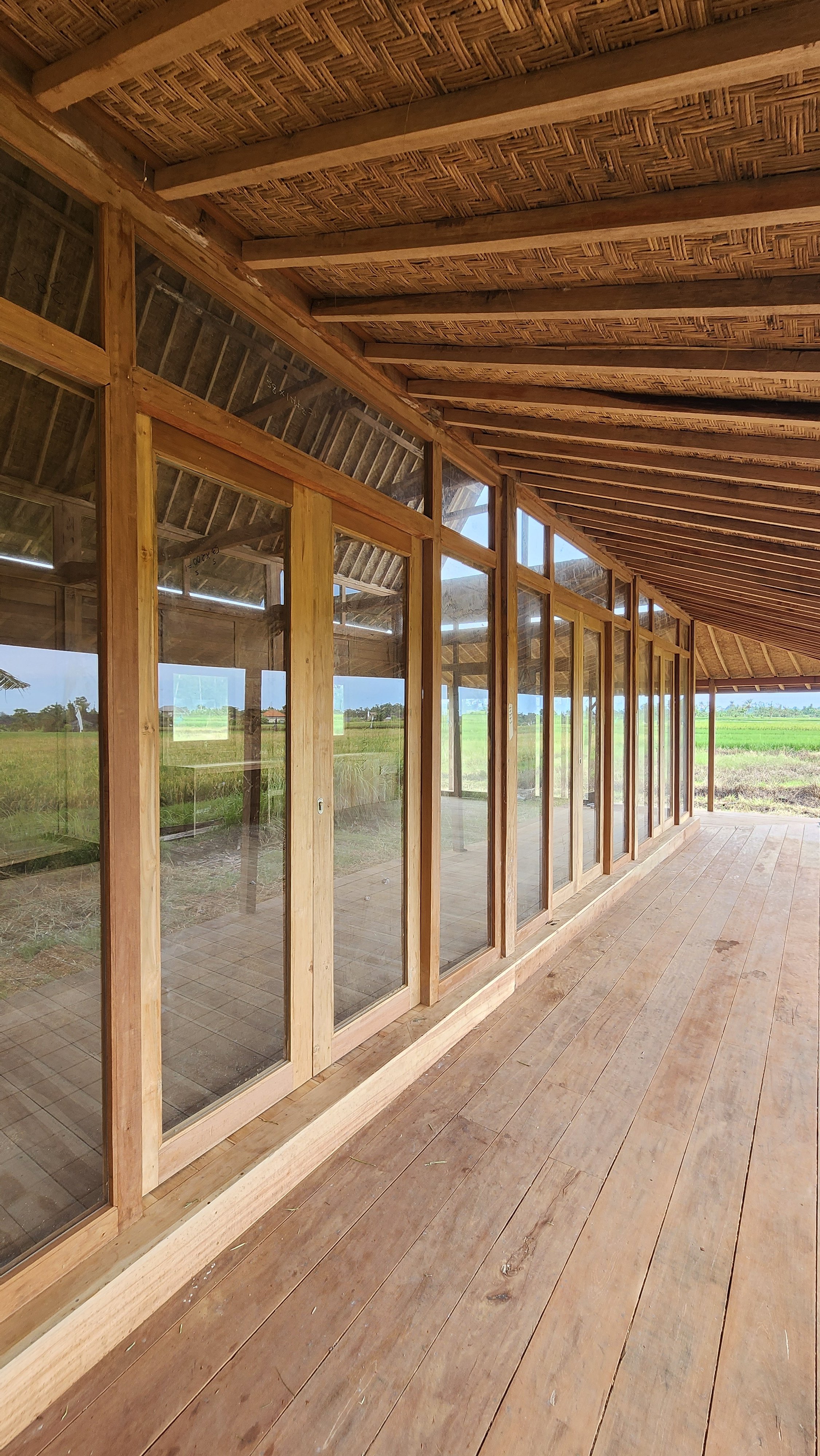 Wooden house with large glass windows overlooking a grassy landscape, featuring a thatched roof and open wooden deck.