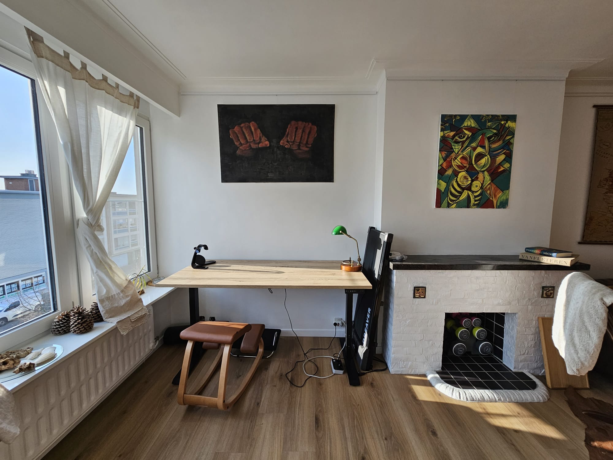 Home office with a wooden desk, chair, artwork on walls, and a decorative fireplace, well-lit by large windows.