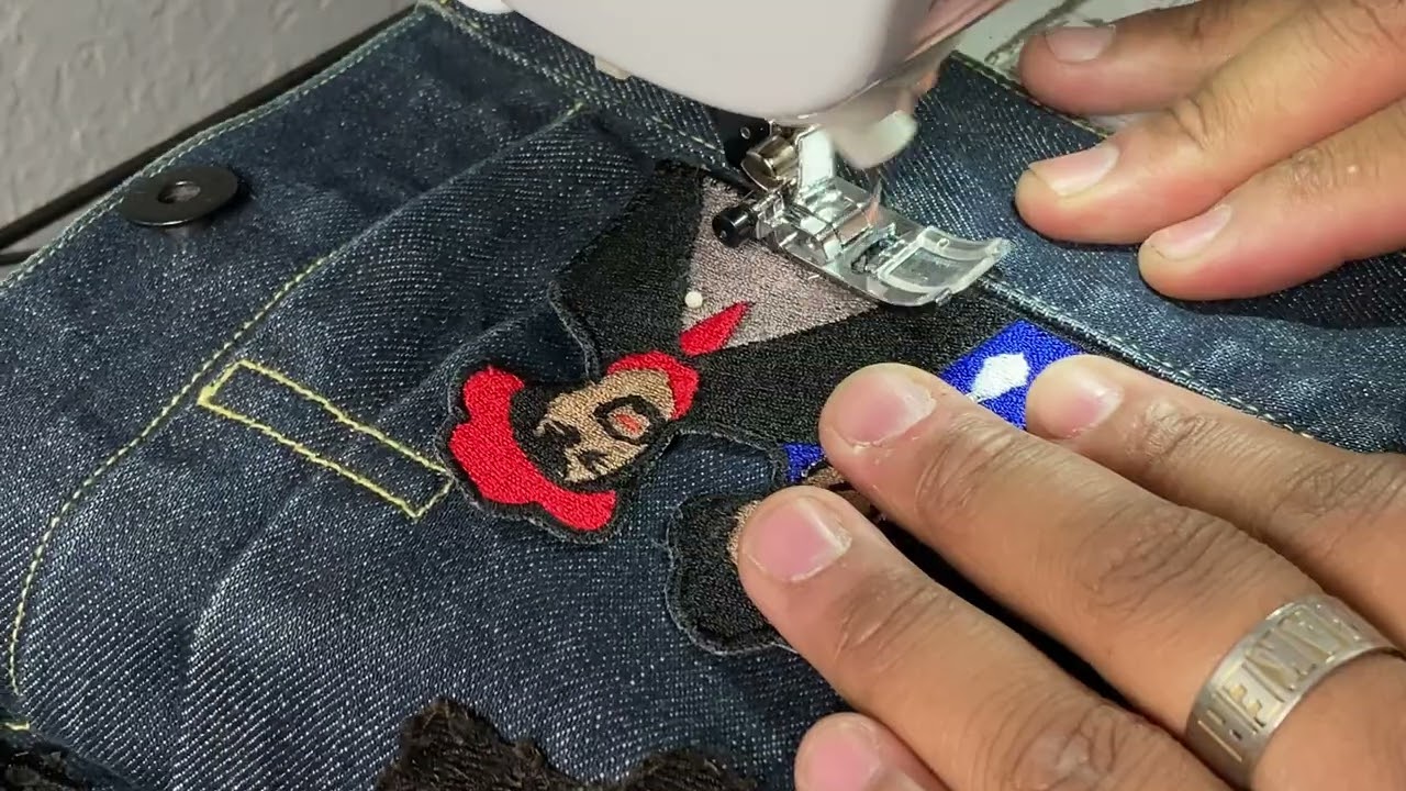 A person using a sewing machine to embroider a design with a red hat and blue shirt on dark fabric, wearing a ring.