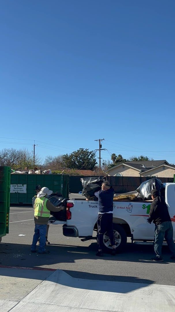 Three people load garbage bags into a white pickup truck near a waste disposal area on a sunny day.