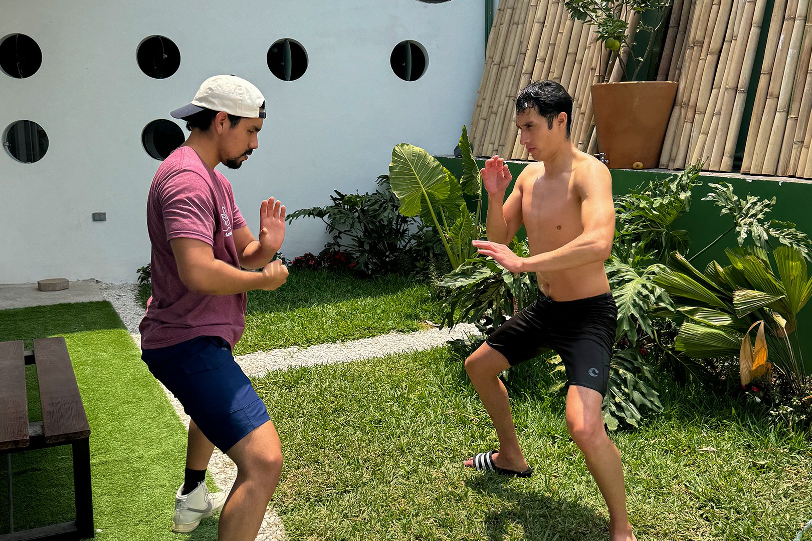 Two men practicing martial arts in a garden, one shirtless and barefoot, the other in a cap and shorts.