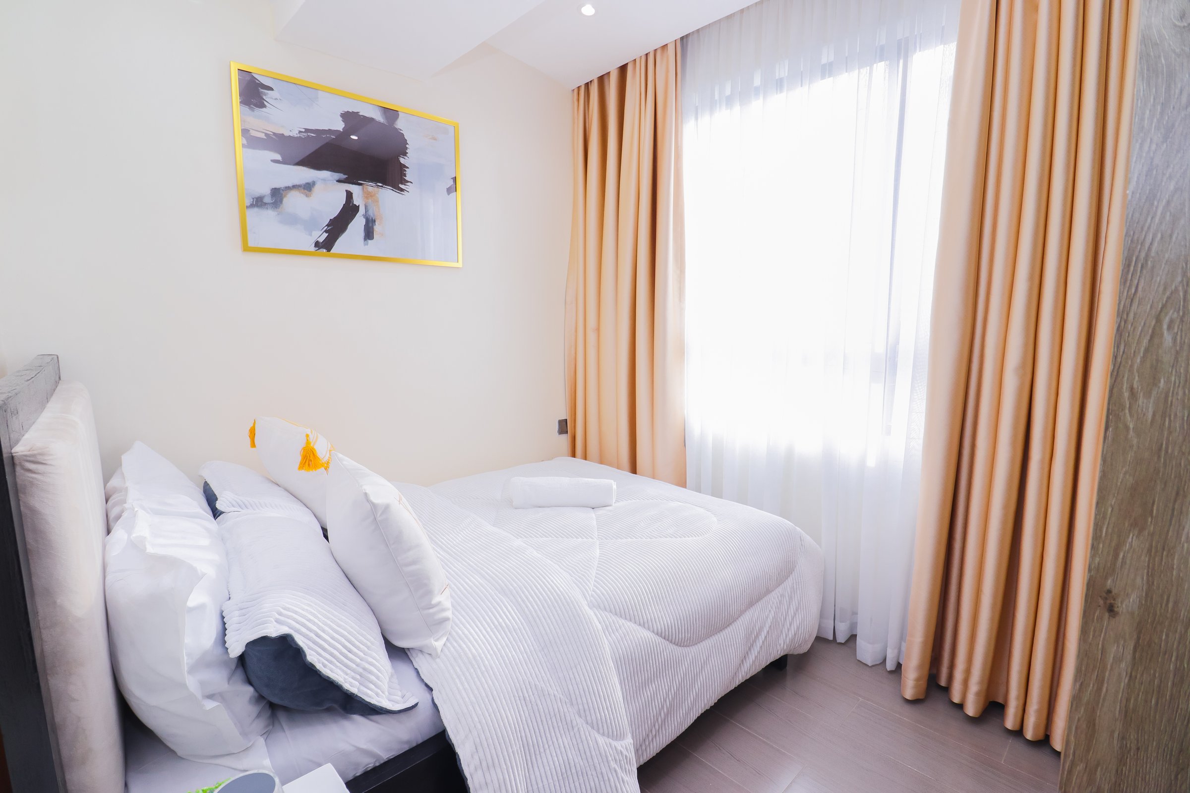 A bright, minimalist bedroom with a neatly made bed, soft lighting, abstract wall art, and floor-to-ceiling curtains.