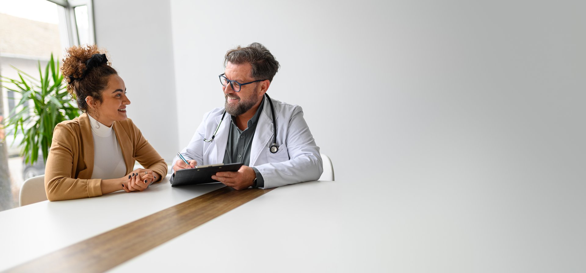 Smiling professional male doctor holding clipboard talking to female patient while sitting at desk in hospital