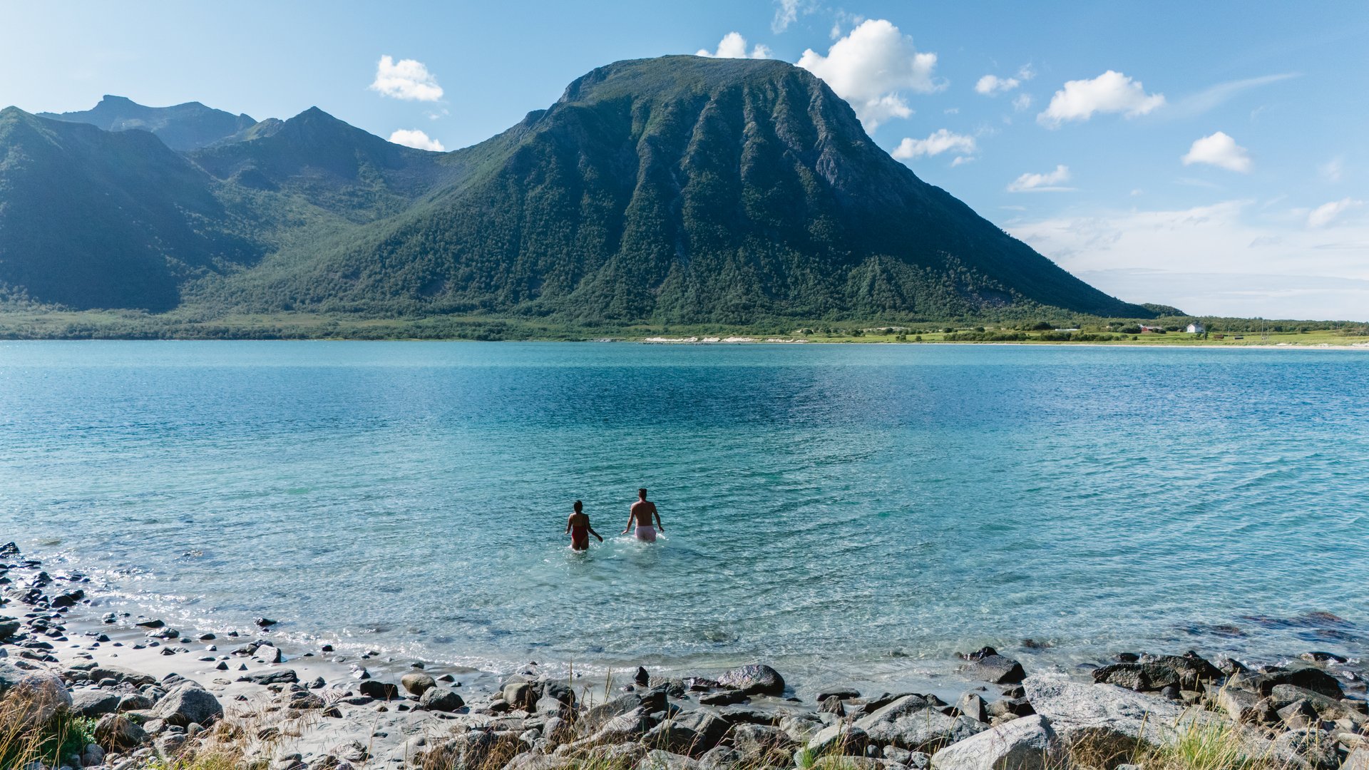 Two individuals wade into the clear blue waters of Lofoten, Norway. Surrounded by majestic mountains and vibrant greenery, this serene summer day invites adventure and relaxation in nature embrace.
