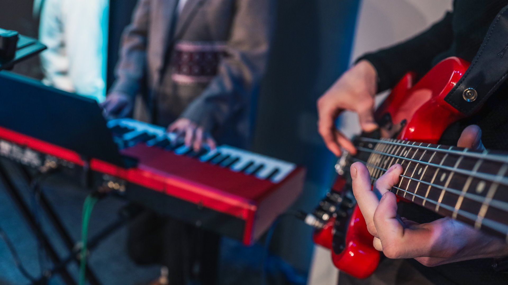 Close-up of hands playing electronic keyboard during live music performance with blurred bassist in background under blue lighting.