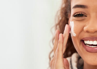Woman smiling while applying cream to her cheek with a blurred background.