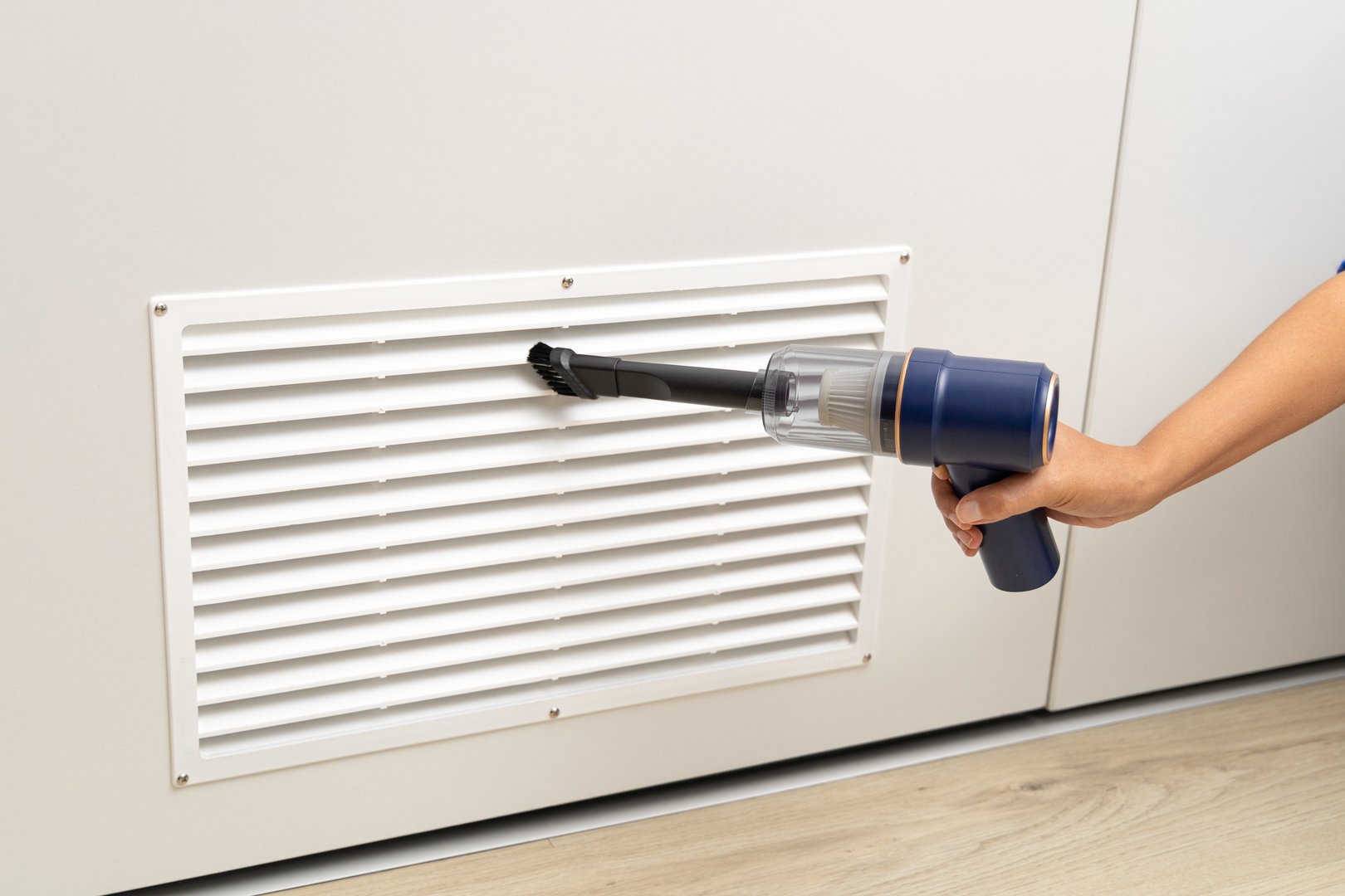 A person is using a vacuum cleaner to clean a vent. The person is holding the vacuum cleaner with both hands and is focused on the task