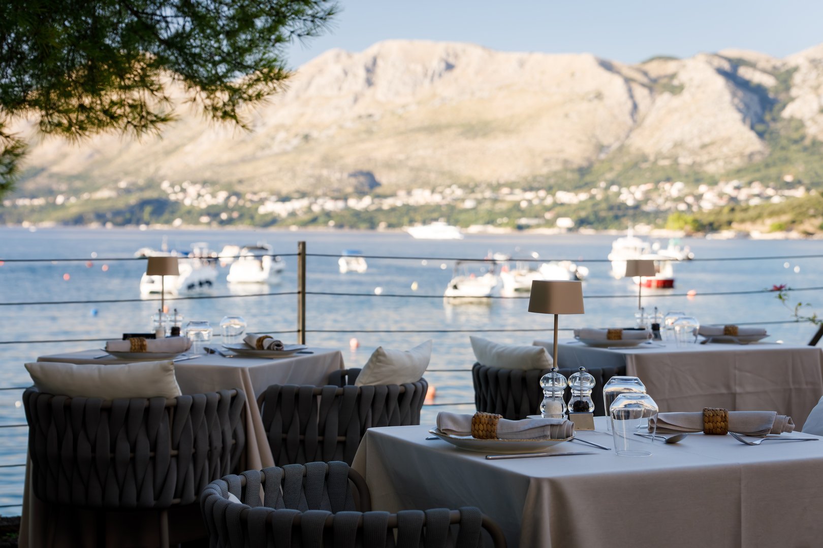 Tables are set for meals at a seaside restaurant, overlooking tranquil waters and distant mountains under a clear blue sky.