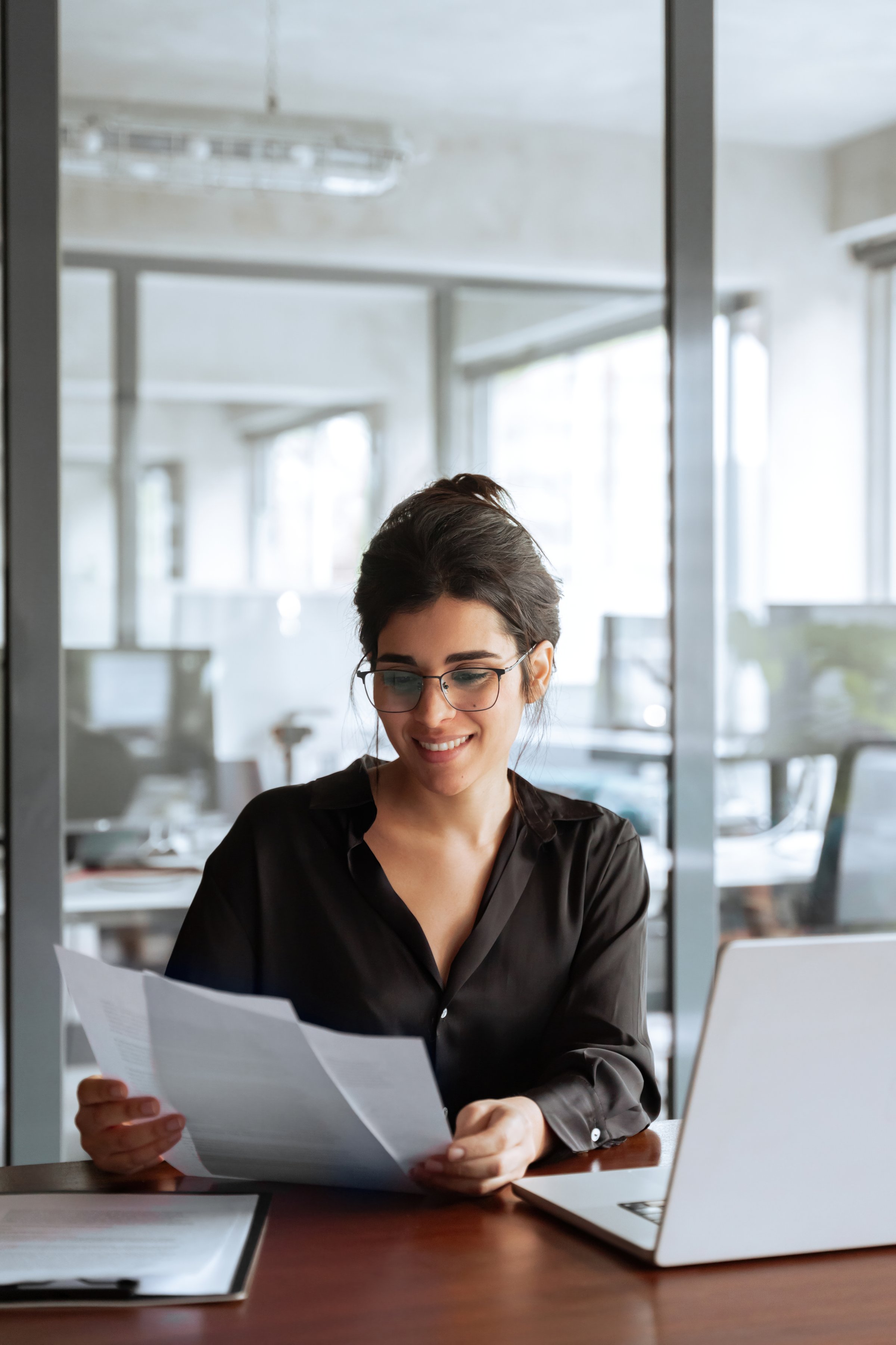 Vertical smiling portrait business woman accountant holding documents, working at laptop pc computer doing online tax invoice, financial report. Latina businesswoman manager with paperwork in office
