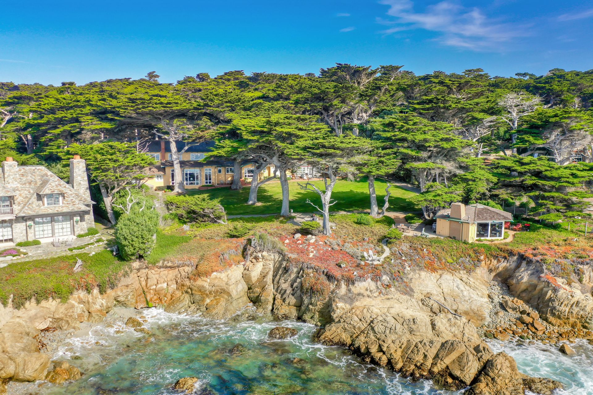 A coastal mansion surrounded by trees perched on rocky cliffs with waves crashing below under a clear blue sky.