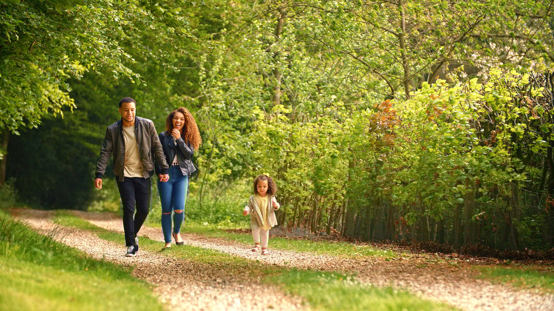 Family With Young Daughter Walking Along Track Through Trees In Countryside Towards Camera Together