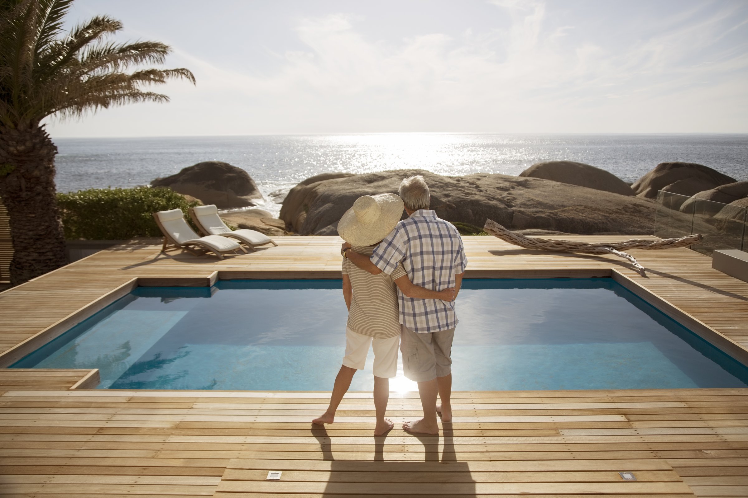 An elderly couple embraces by a pool on a wooden deck, overlooking the ocean with large rocks and a sunset in the background.