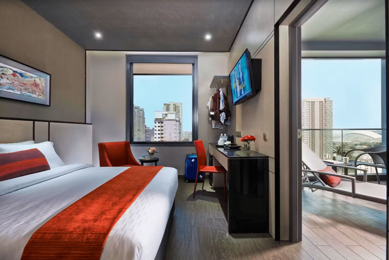 Modern hotel room with bed, TV, desk, and balcony overlooking cityscape. Red accents on bed and chairs.