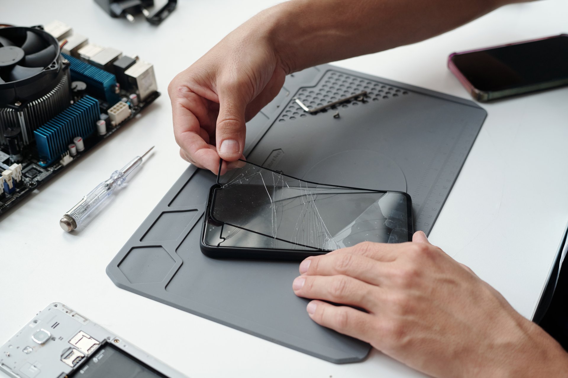 Close-up of hands meticulously repairing cracked smartphone screen, with disassembled phone parts and tools on workbench. Visible devices include screwdriver, smartphone, and motherboard