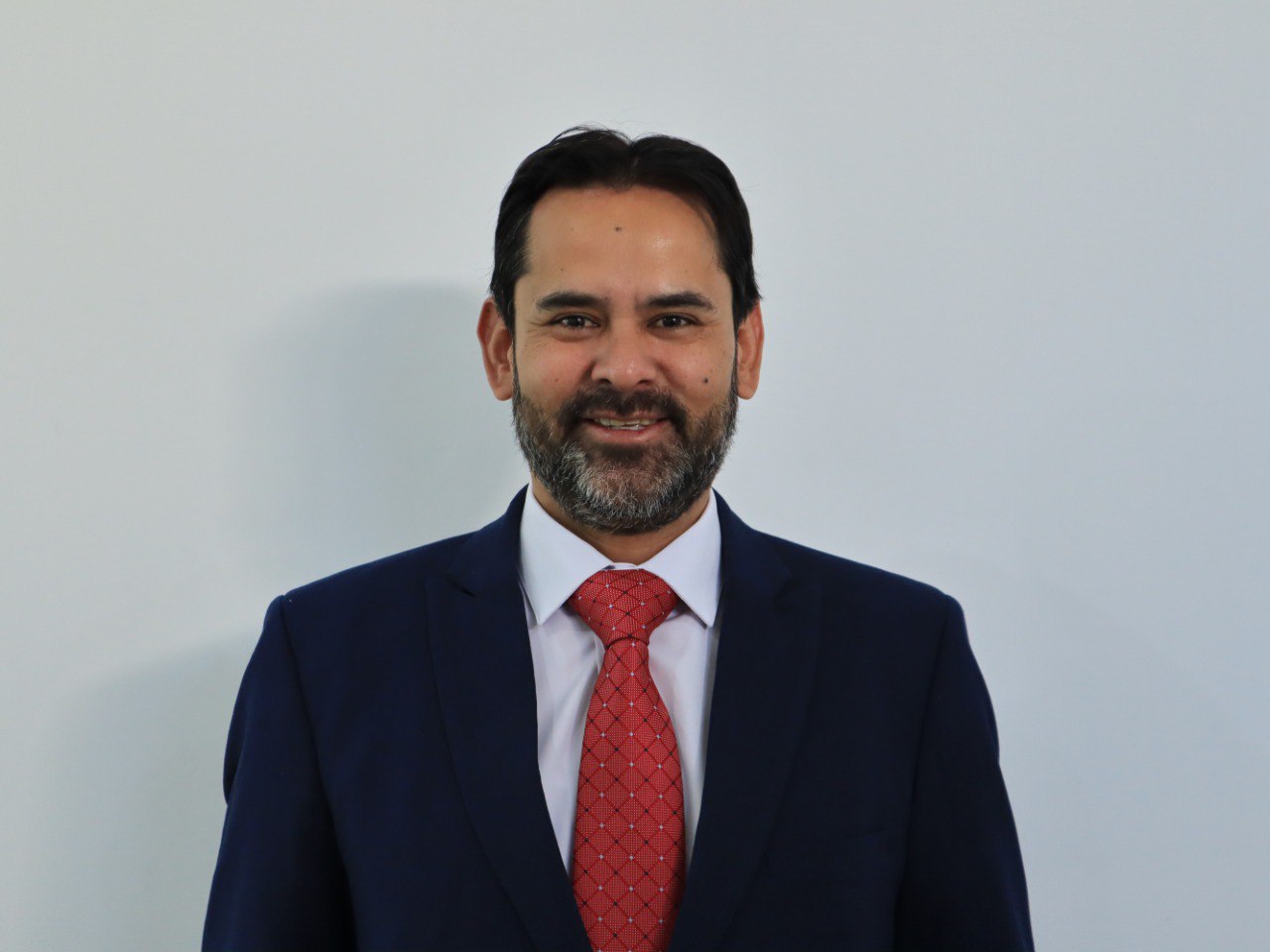 Smiling man in a suit with a red tie against a plain background.