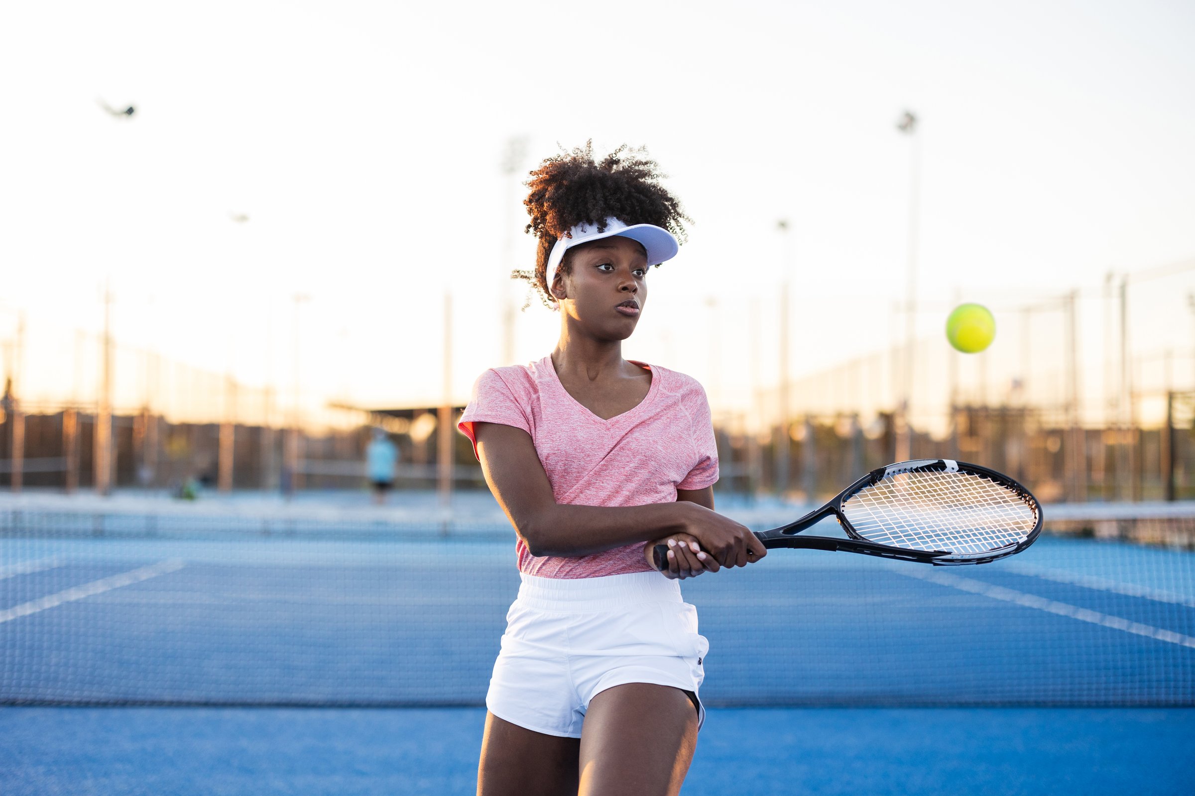 Young black woman wearing a pink shirt and white shorts and visor, holding a tennis racket, prepares to hit a ball on a blue tennis court at sunset