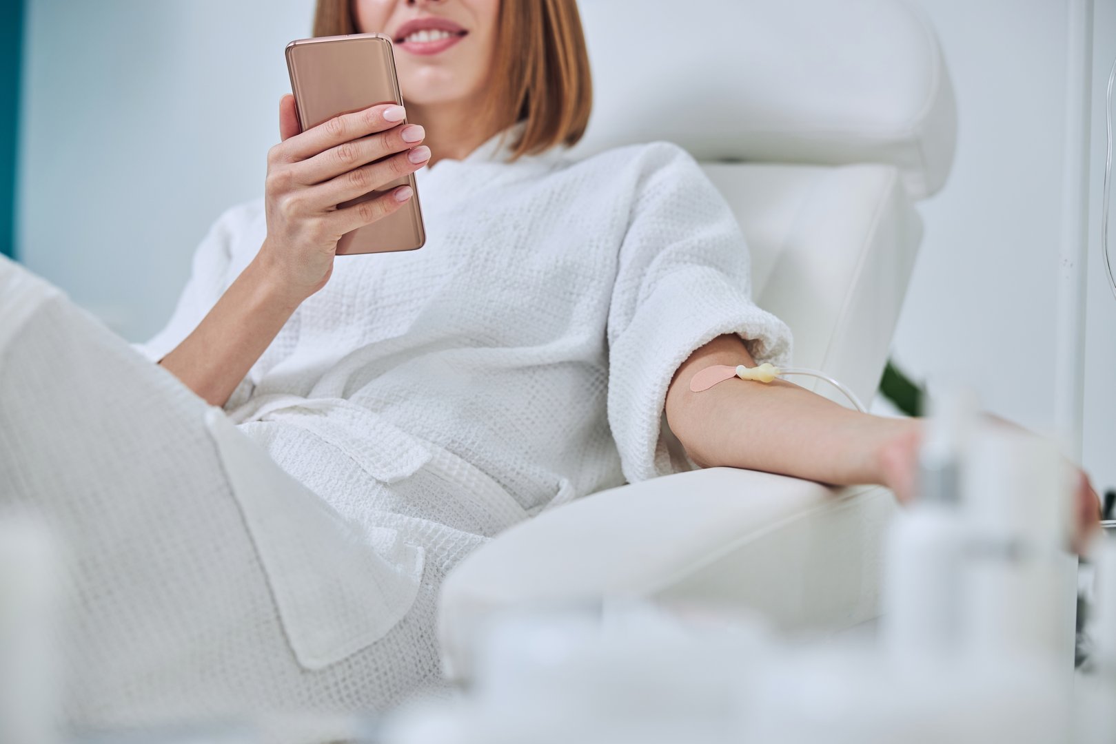 Cropped head portrait of unrecognized young lady in white bathrobe holding mobile phone in hand while receiving intravenous vitamin drip treatment in wellness center