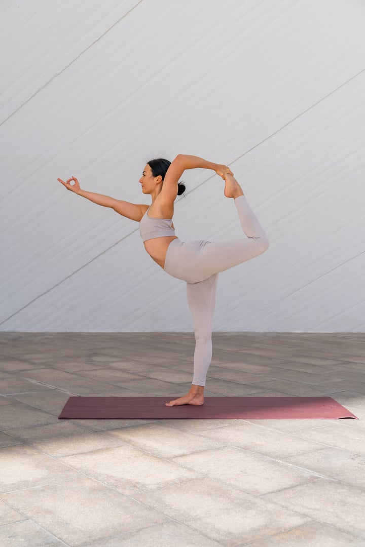 Brunette woman dressed in sportswear performing Natarajasana yoga pose or dancer's pose on modern, minimalist background.