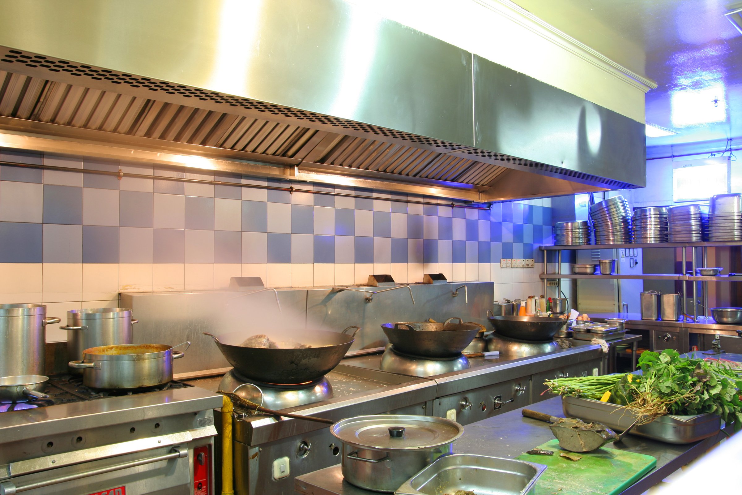 Professional kitchen with woks on stoves, steam rising, stainless steel surfaces, and fresh greens on the counter.