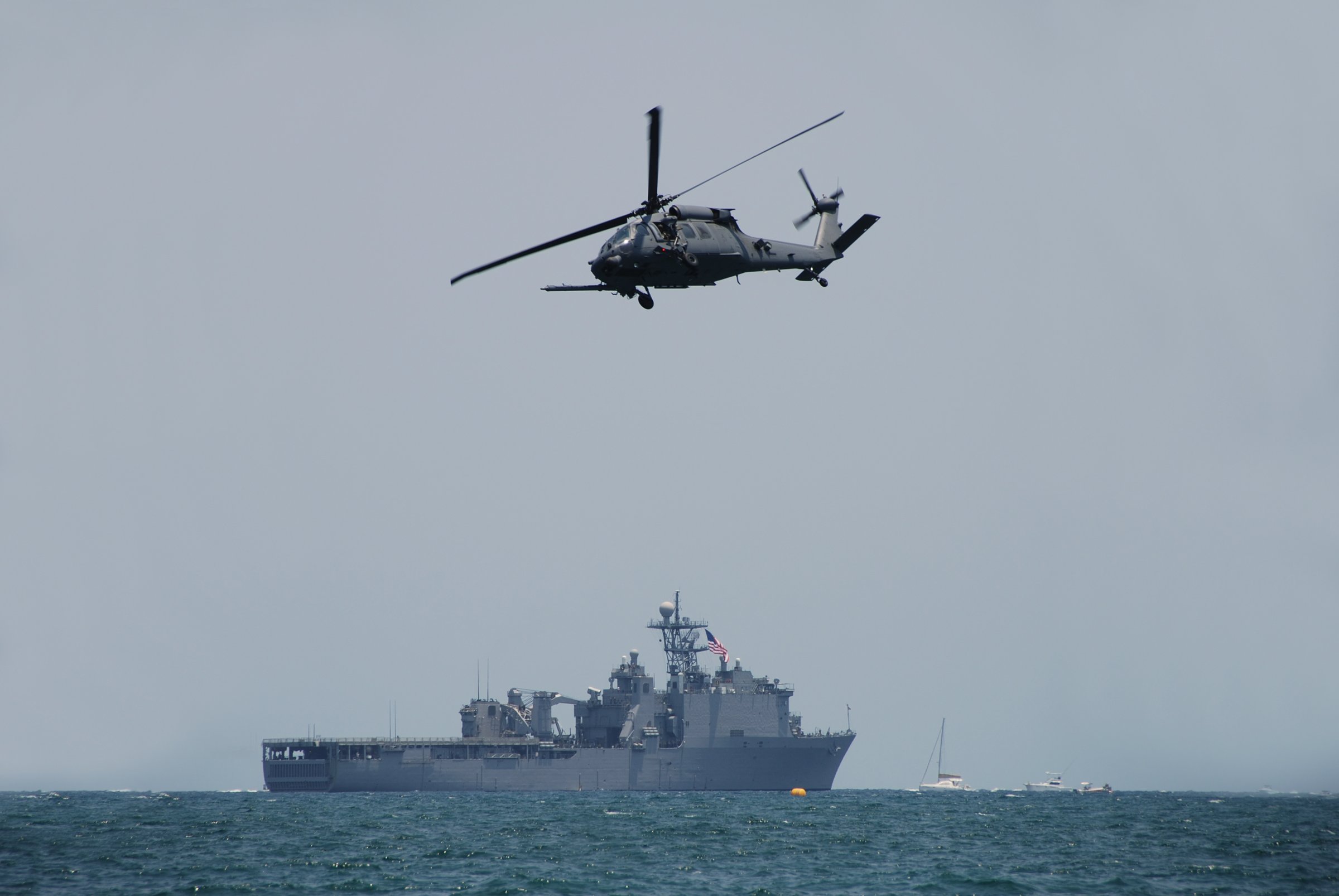 Helicopter flying over the ocean with a naval ship in the background. The sky is overcast and the water appears calm.