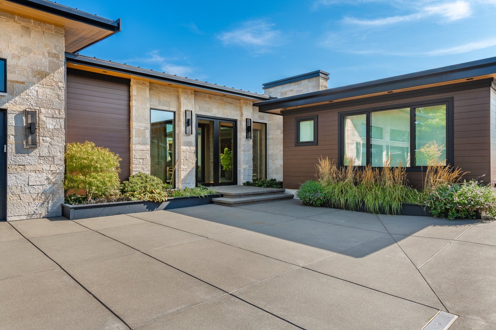 Exterior photograph of a mountain modern home residence a bright blue sky summer day and the house has an expansive ocean front deck with a swimming pool and a hot tub the tide is low and there are coastal views we also see an enclosed outdoor living area with stainless steel cabinets cooking facilities dining area wooden furniture the front facade of the home shows a flat roof and two large garage doors with glass windows the home has stone and wood cladding waterfront deck leads to modern living room through floor to ceiling glass doors