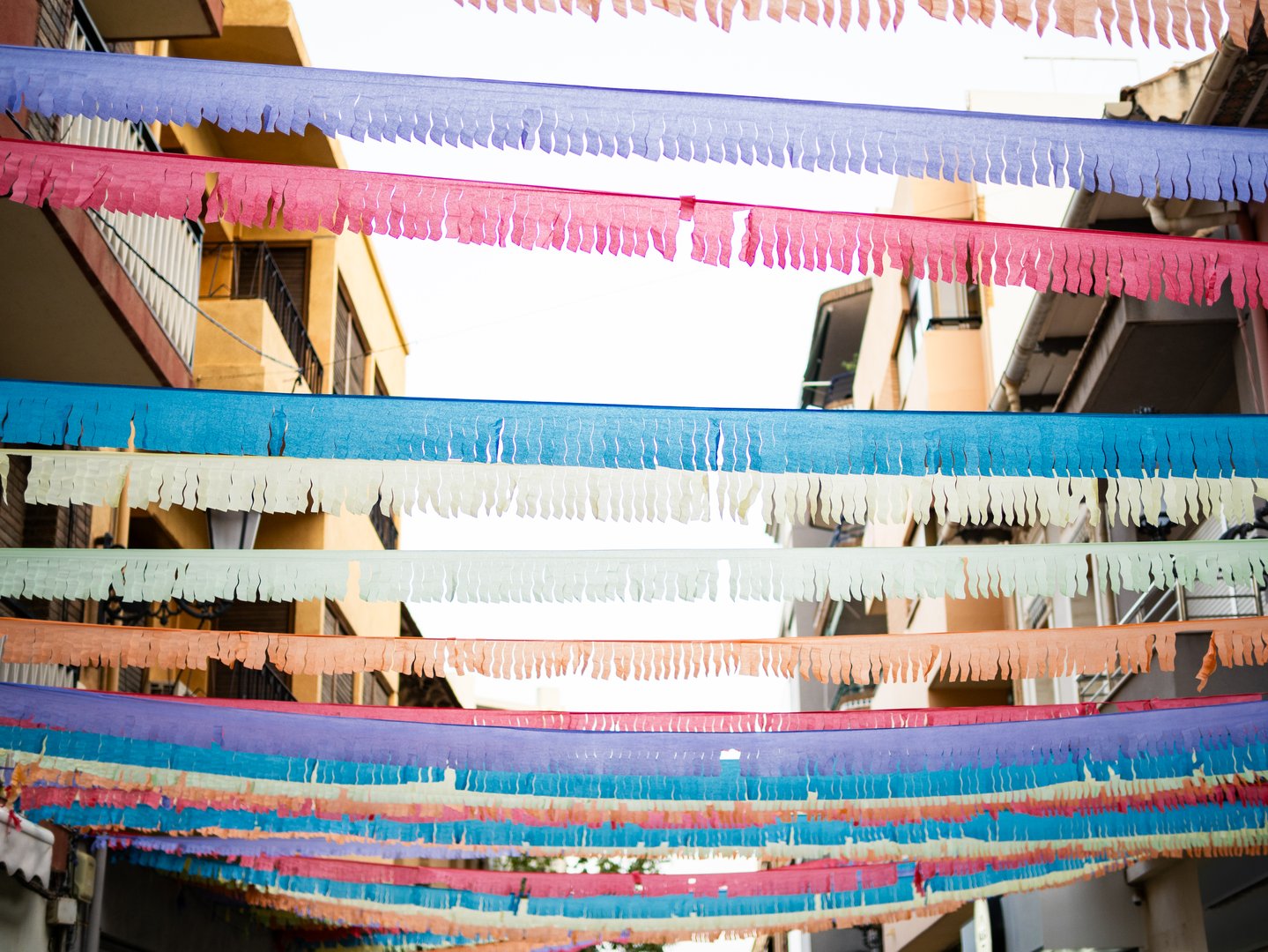 Brightly colored paper garland streamers hang overhead in a lively street. The decorations create a cheerful ambiance as people prepare for a local celebration during daylight.