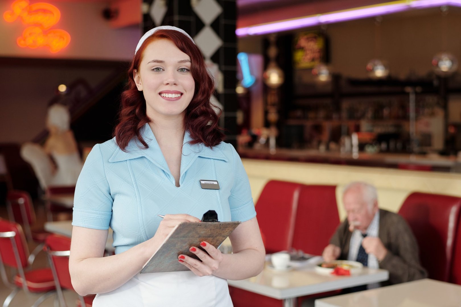 Smiling waitress holding a notepad in 1950s style diner with red upholstery and elderly man eating at table in background creating nostalgic setting